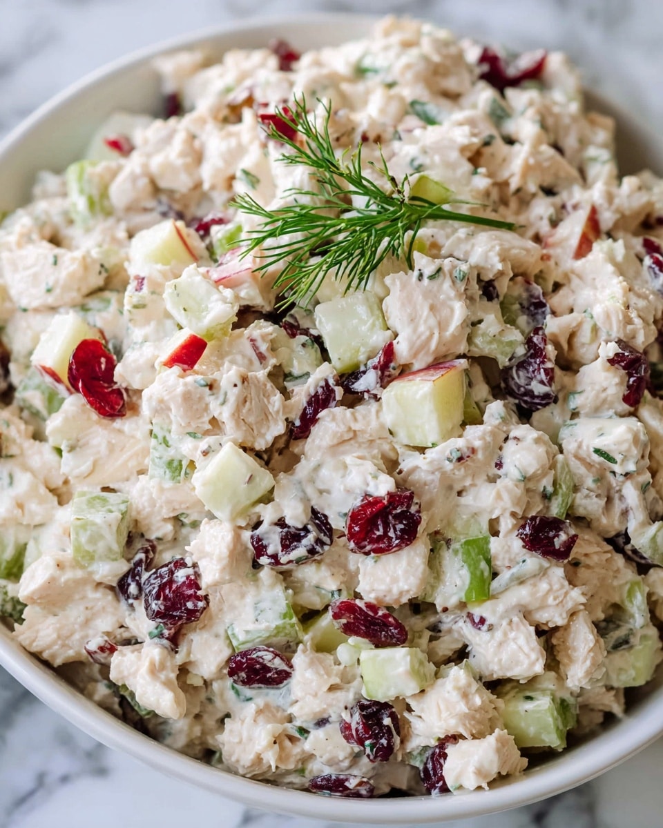 A close-up view of a bowl filled with creamy chicken salad that has multiple layers of tiny chopped pieces. The salad is creamy white with chunks of light beige chicken, green celery bits, small cubes of pale yellow apple, dark red dried cranberries, and red apple pieces mixed throughout. The salad texture looks soft and chunky with a few fresh green sprigs on top for decoration. The bowl is white, and it sits on a surface with a white marbled texture. photo taken with an iphone --ar 4:5 --v 7