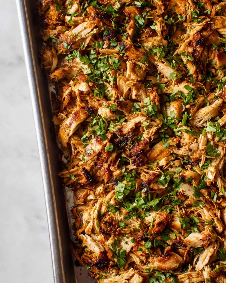 The image shows a close-up of a baking tray filled with shredded cooked chicken that has a golden-brown grilled texture with some charred edges. The cooked chicken pieces are unevenly spread out, creating a layered effect with strands visible throughout. Bright green chopped herbs are sprinkled on top, providing a fresh contrast to the warm brown tones of the chicken. The tray is placed on a white marbled surface. photo taken with an iphone --ar 4:5 --v 7