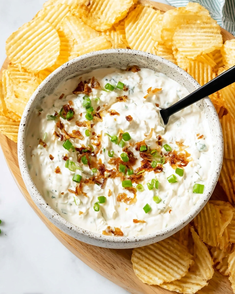 A white speckled bowl filled with a creamy white dip that has small bits of browned onions and is topped with scattered bright green chopped scallions, placed on a light wooden board surrounded by ridged, pale yellow potato chips, with a black spoon dipping into the creamy mixture, all set on a white marbled surface. photo taken with an iphone --ar 4:5 --v 7