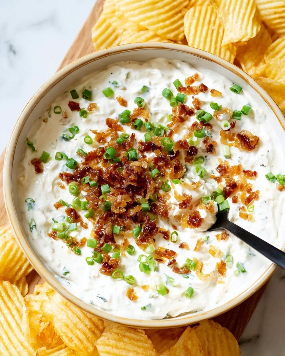 A close-up of a creamy white dip in a beige rimmed bowl, topped with small pieces of browned caramelized onions and finely chopped green onions scattered evenly across the surface, creating a mix of golden brown and bright green colors on the white dip base; a black spoon rests partially submerged on the right side, and the bowl is surrounded by ridged yellow potato chips, all placed on a white marbled surface with a wooden board partially visible underneath. photo taken with an iphone --ar 4:5 --v 7