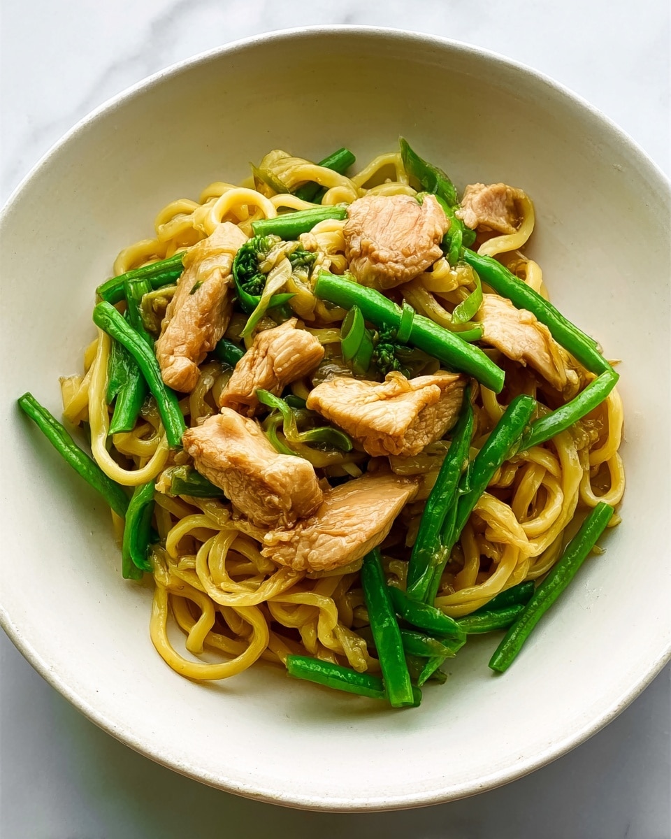A white bowl holds a serving of thick yellow noodles mixed with pieces of light brown cooked chicken and bright green fresh green beans. The chicken pieces are cut into small, uneven strips with a slightly browned and juicy texture. The green beans are whole, crisp, and placed evenly among the noodles. The noodles are curled and soft, sitting at the base and mixed throughout the dish. The bowl is set on a white marbled surface. photo taken with an iphone --ar 4:5 --v 7
