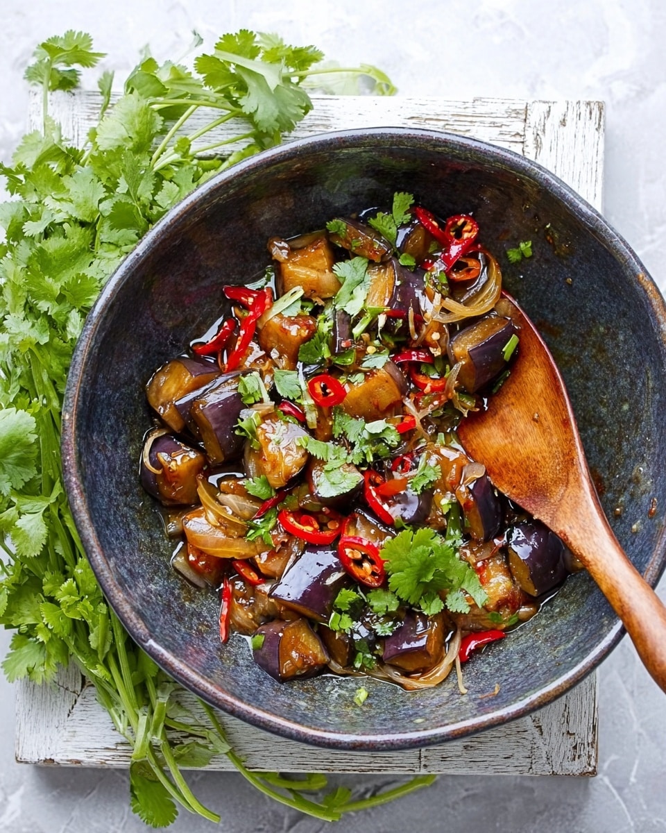 The image shows a dark grey rustic bowl filled with a colorful dish. Inside, there are brown pieces of cooked eggplant with soft texture and a shiny sauce that looks glossy. Bright red chili slices and green coriander leaves are scattered throughout, adding fresh color. There are some translucent, cooked onion slices mixed in too. A wooden spoon with a long handle rests inside the bowl. The bowl sits on a rough white-painted wooden surface with fresh green coriander bunch beside it, all set against a white marbled background. photo taken with an iphone --ar 4:5 --v 7