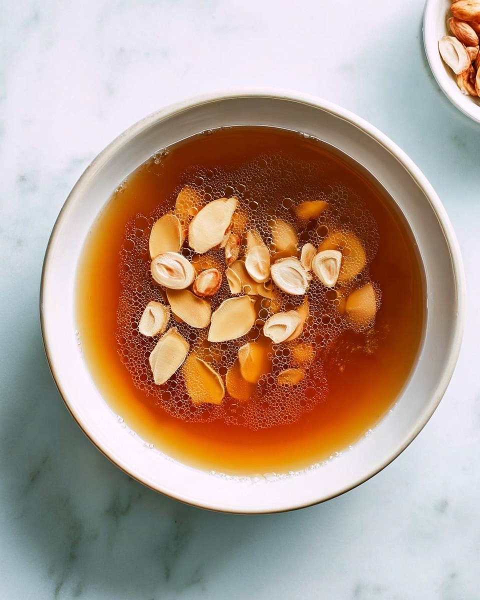 A white bowl filled with a clear brown broth, floating on the surface are several small, light brown, sliced garlic pieces and some unpeeled thinly sliced garlic chips. The broth has tiny bubbles around the edges, creating a fresh and warm look. The bowl sits on a white marbled surface, giving a clean and bright setting. Photo taken with an iphone --ar 4:5 --v 7