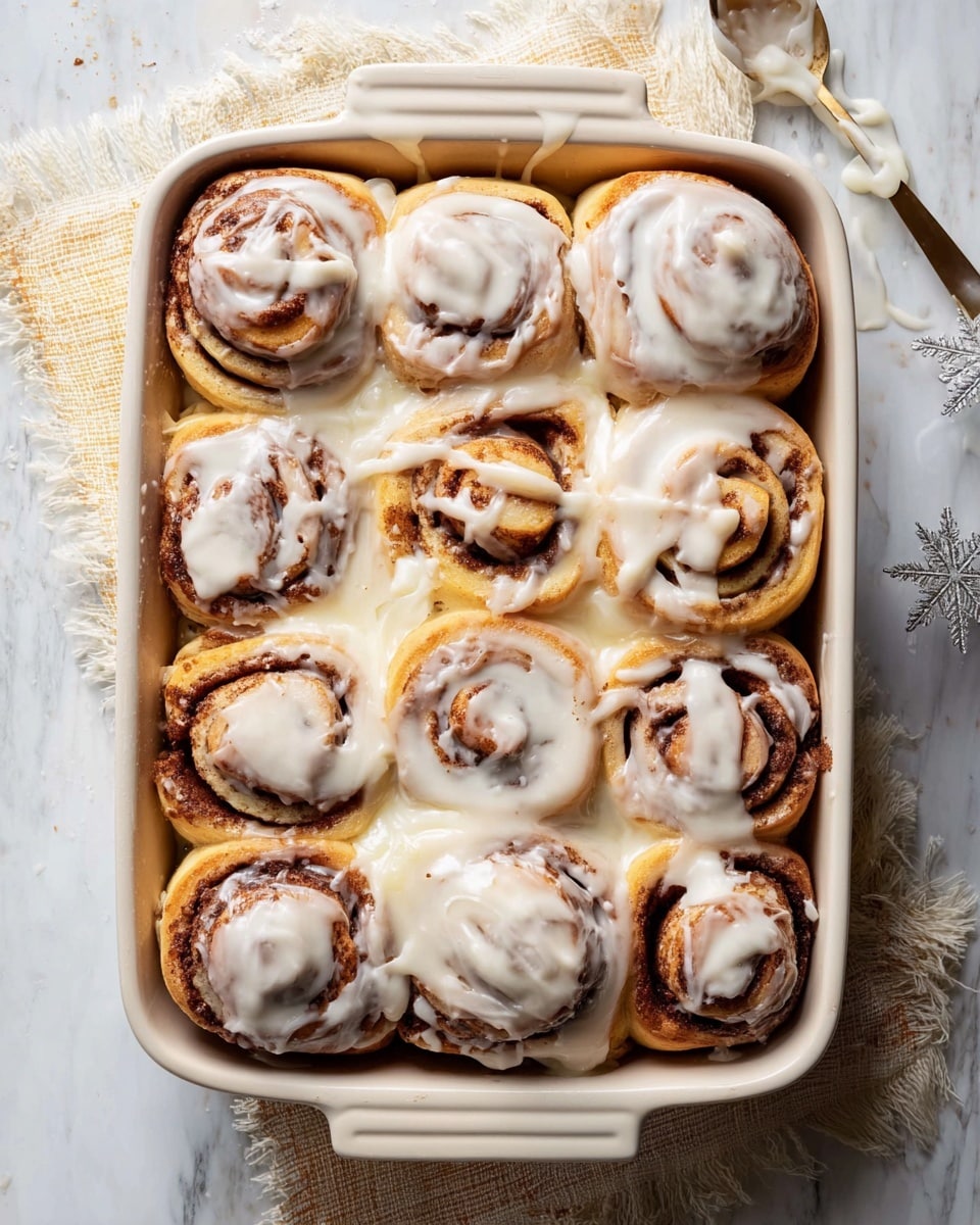 A white rectangular baking dish holds twelve cinnamon rolls arranged in a 4 by 3 grid, each roll showing distinct layers of soft golden-brown dough spiraled with dark cinnamon filling. The rolls are covered generously with thick, creamy white icing that drips down the sides and pools slightly around the edges. Some rolls have more icing on top, creating a shiny, smooth texture, while the dough beneath reveals a slightly crispy, browned surface. The dish sits on a white marbled surface with a cloth underneath, and a spoon with some icing is placed nearby. Photo taken with an iphone --ar 4:5 --v 7