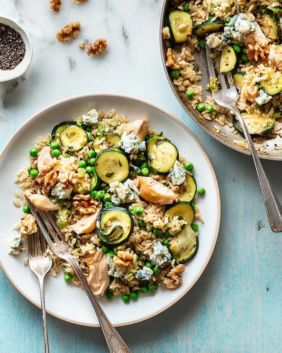 A white plate filled with a mixed rice dish on a white marbled surface. The dish has three main layers: at the bottom, light brown cooked rice, scattered green peas and small chunks of cooked white chicken meat. On top of this, sliced cooked green zucchini rounds and slices of sautéed onions are spread evenly. The top layer is decorated with small pieces of creamy blue cheese and walnut halves. A silver fork rests on the left side of the plate. Next to it, there is a silver pan with more of the same dish, with a silver spoon resting inside. In the background near the top, a small white bowl holds mixed black and white peppercorns. photo taken with an iphone --ar 4:5 --v 7