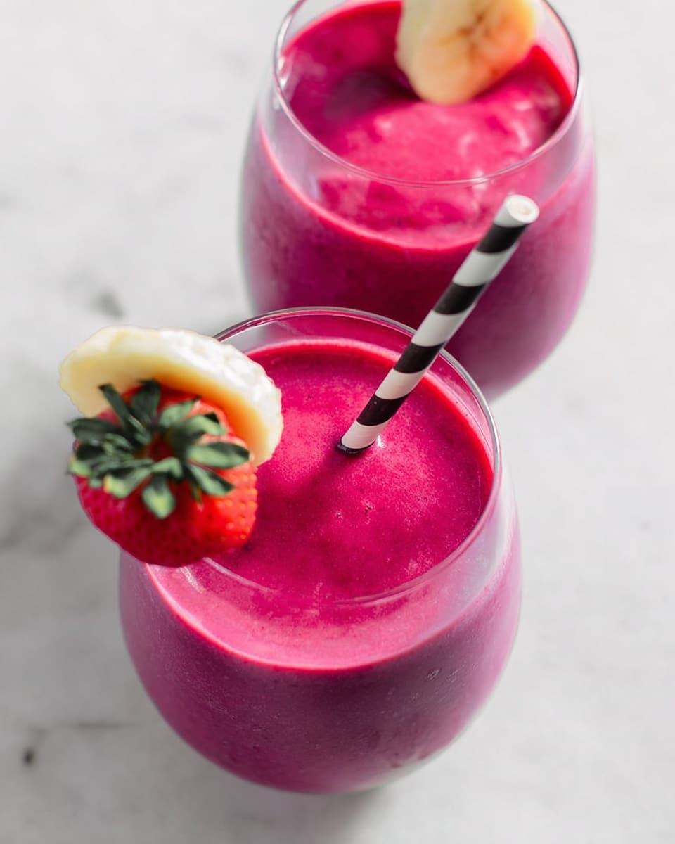 The image shows two clear glasses filled with a thick, bright magenta smoothie with a smooth, creamy texture on top. Each glass has a black and white striped straw placed slightly off-center. The glass in the back is garnished with a slice of light yellow banana and a whole red strawberry with green leaves, placed on the rim. The scene is set on a white marbled surface with soft lighting highlighting the vibrant colors of the drink and fruit. photo taken with an iphone --ar 4:5 --v 7