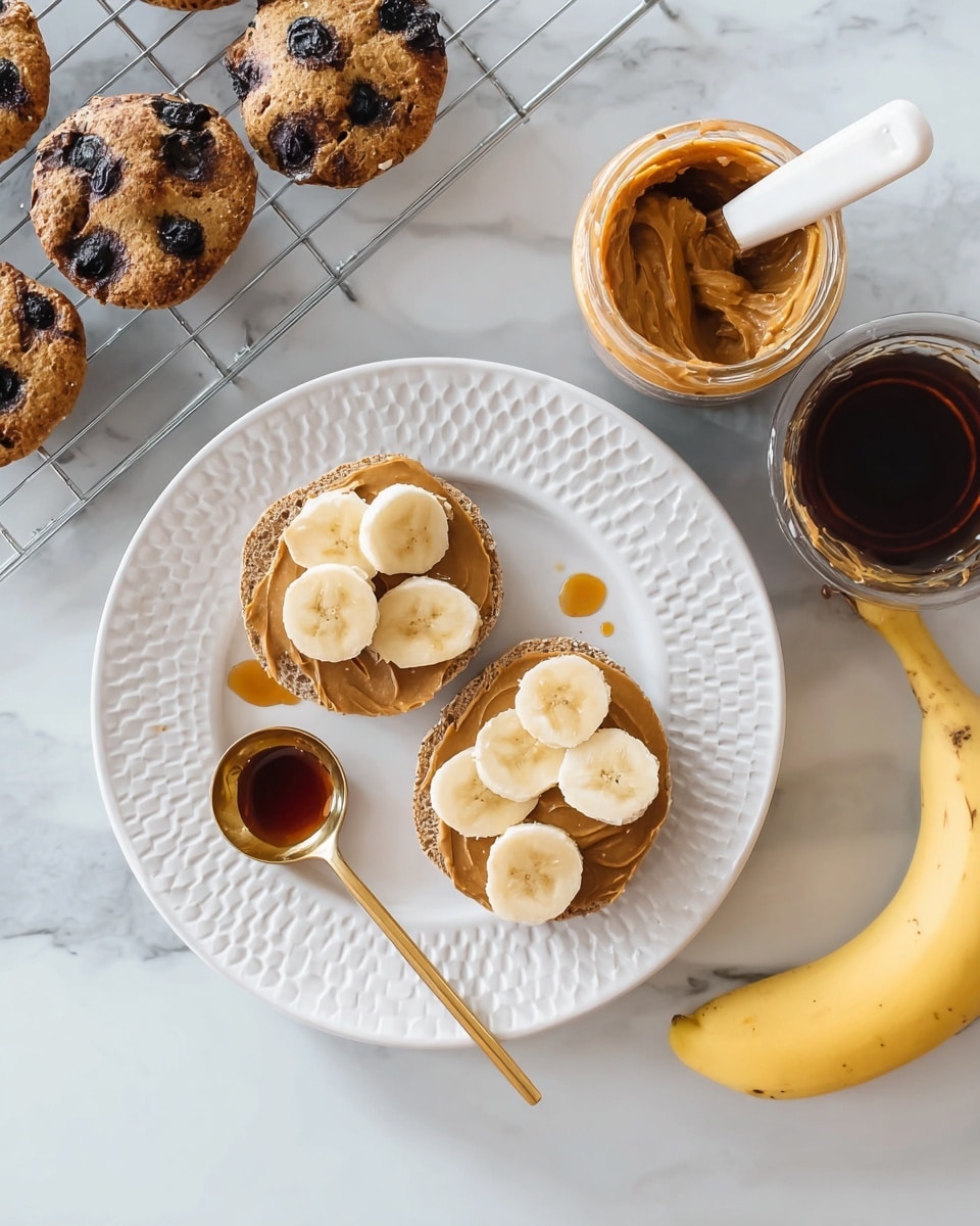 The image shows two halves of a donut-shaped blueberry muffin placed on a white plate with delicate swirl patterns. Each muffin half is topped with a light brown layer of nut butter and covered by four or five slices of fresh yellow banana arranged evenly. A golden measuring spoon holding a dark amber syrup is on the plate next to the muffin halves. Around the plate, there is an open jar of nut butter with a knife inside, a jar of syrup with the lid off, a partly peeled ripe banana, and a cooling rack holding three more whole blueberry muffins. The scene is set on a white marbled surface. Photo taken with an iphone --ar 4:5 --v 7