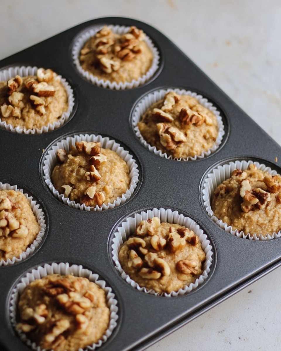 The image shows a dark gray metal muffin tray with twelve muffins in white paper liners, each filled with light brown batter. On top of each muffin batter is a piece of walnut, giving a textured look with uneven shapes and light brown tones. The tray is placed on a white marbled surface, and the lighting highlights the soft, slightly wet texture of the batter and the contrast of the nuts on top. photo taken with an iphone --ar 4:5 --v 7