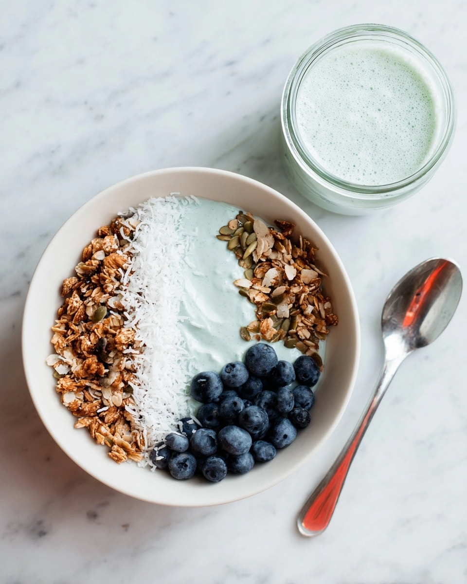 A white bowl on a white marbled surface holds a breakfast dish with four distinct parts: pale blue yogurt with a slightly bumpy texture takes up half the bowl, next to it is a line of white shredded coconut, followed by crunchy brown granola with visible seeds and nuts, and a small pile of fresh dark blue blueberries at the bottom right. Next to the bowl is a small glass jar filled with the same pale blue yogurt. A silver spoon lies to the right of the bowl, reflecting some red color from the surroundings. photo taken with an iphone --ar 4:5 --v 7