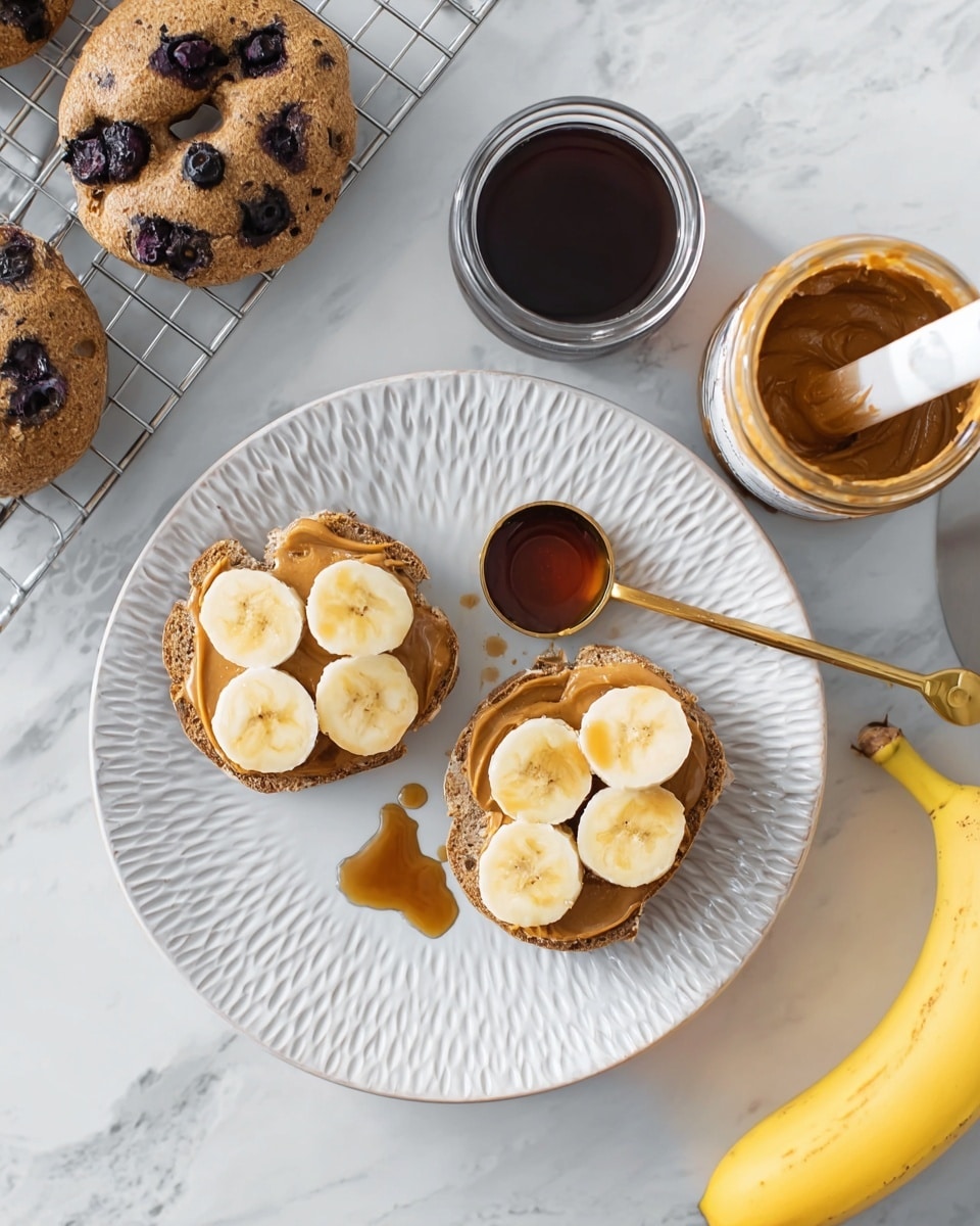 The image shows two blueberry bagel halves on a white plate with a textured swirl pattern. Each bagel half has a thick spread of peanut butter and is topped with four to five round banana slices, some of which are slightly browned. A brass measuring spoon with dark syrup rests on the plate, with a small drop of syrup spilled next to it. To the right, an open jar of smooth peanut butter with a white-handled knife inside is visible, along with a glass jar of dark syrup. Below the jars, a partially peeled ripe banana rests on a white marbled surface. In the top left corner, three whole blueberry bagels cool on a metal rack. The photo taken with an iphone --ar 4:5 --v 7