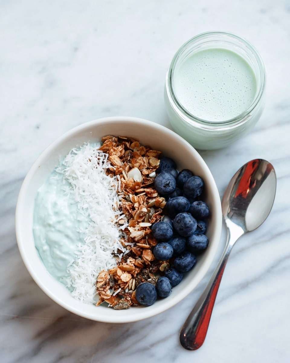 A white bowl shows a layered smoothie bowl with four distinct sections: a smooth pale blue-green smoothie on the left bottom, topped with a neat line of white shredded coconut next to a thick layer of mixed granola with seeds and nuts on the top right, and a small cluster of fresh dark blue blueberries placed at the bottom right. Beside the bowl sits a matching jar of the same blue-green smoothie and a shiny spoon on a white marbled surface. photo taken with an iphone --ar 4:5 --v 7