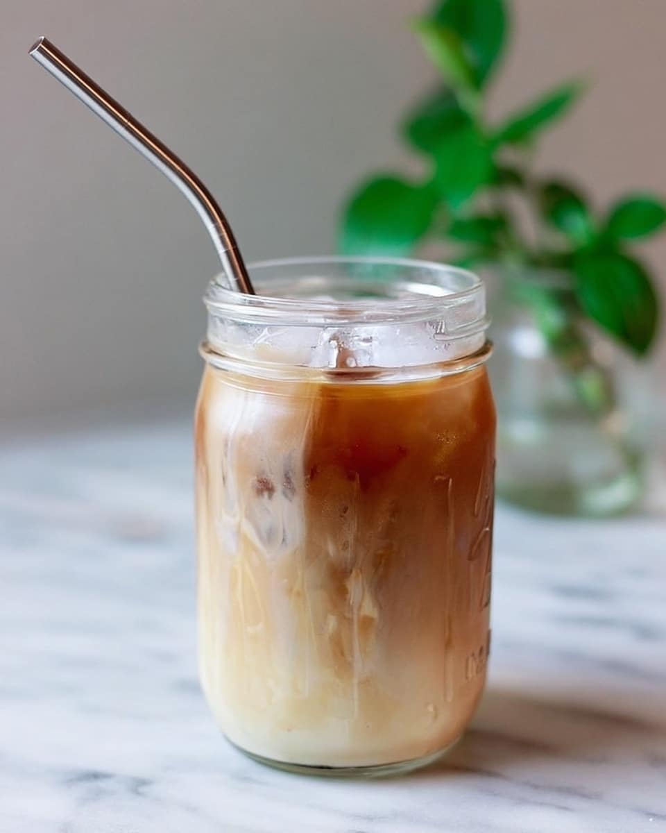 A clear glass jar filled with a cold drink showing three visible layers: a white milky layer at the bottom, a light brown coffee layer in the middle, and a creamy frothy layer on top. The jar contains ice cubes and a metal straw leaning against the side. The background is a blurred white marbled texture with some green leaves visible behind. Photo taken with an iphone --ar 4:5 --v 7