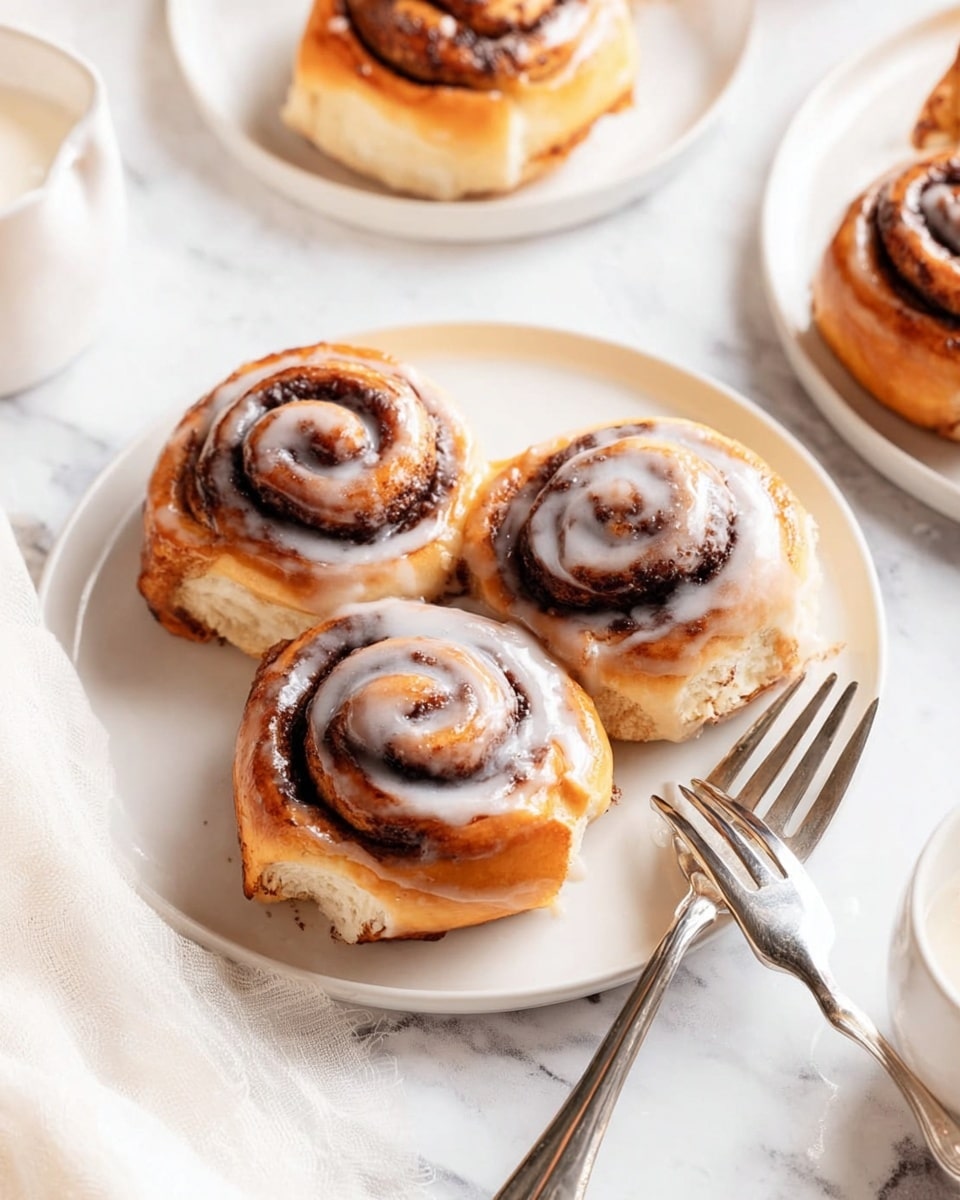The image shows three cinnamon rolls on a white round plate placed on a white marbled surface. Each cinnamon roll has a golden-brown baked dough base with a dark brown spiral of cinnamon filling visible on top, lightly drizzled with white icing that adds a glossy texture. The rolls are fluffy and soft with a slightly crisp outer edge. Two silver forks rest on the right side of the plate. Part of a white plate holding more cinnamon rolls and a white cloth napkin are also visible in the scene. Photo taken with an iphone --ar 4:5 --v 7