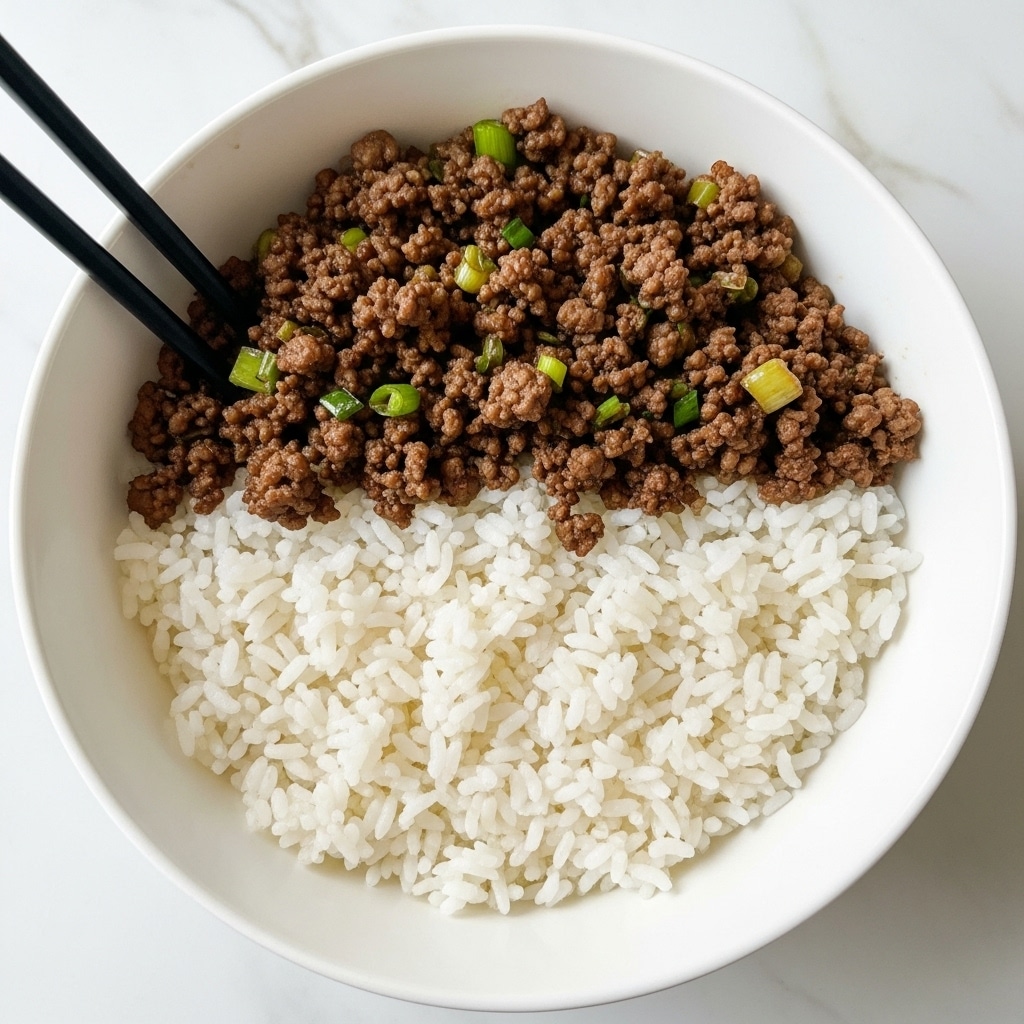 A white bowl filled with a base layer of fluffy white rice, topped with a generous layer of dark brown minced beef mixed with small pieces of green onions, giving a slightly fresh contrast. The beef looks crispy and well-cooked with bits of green scattered throughout. On the left side of the bowl, a pair of black chopsticks rests angled inside the bowl. The setting is on a white marbled surface with soft natural light highlighting the textures and colors. photo taken with an iphone --ar 4:5 --v 7