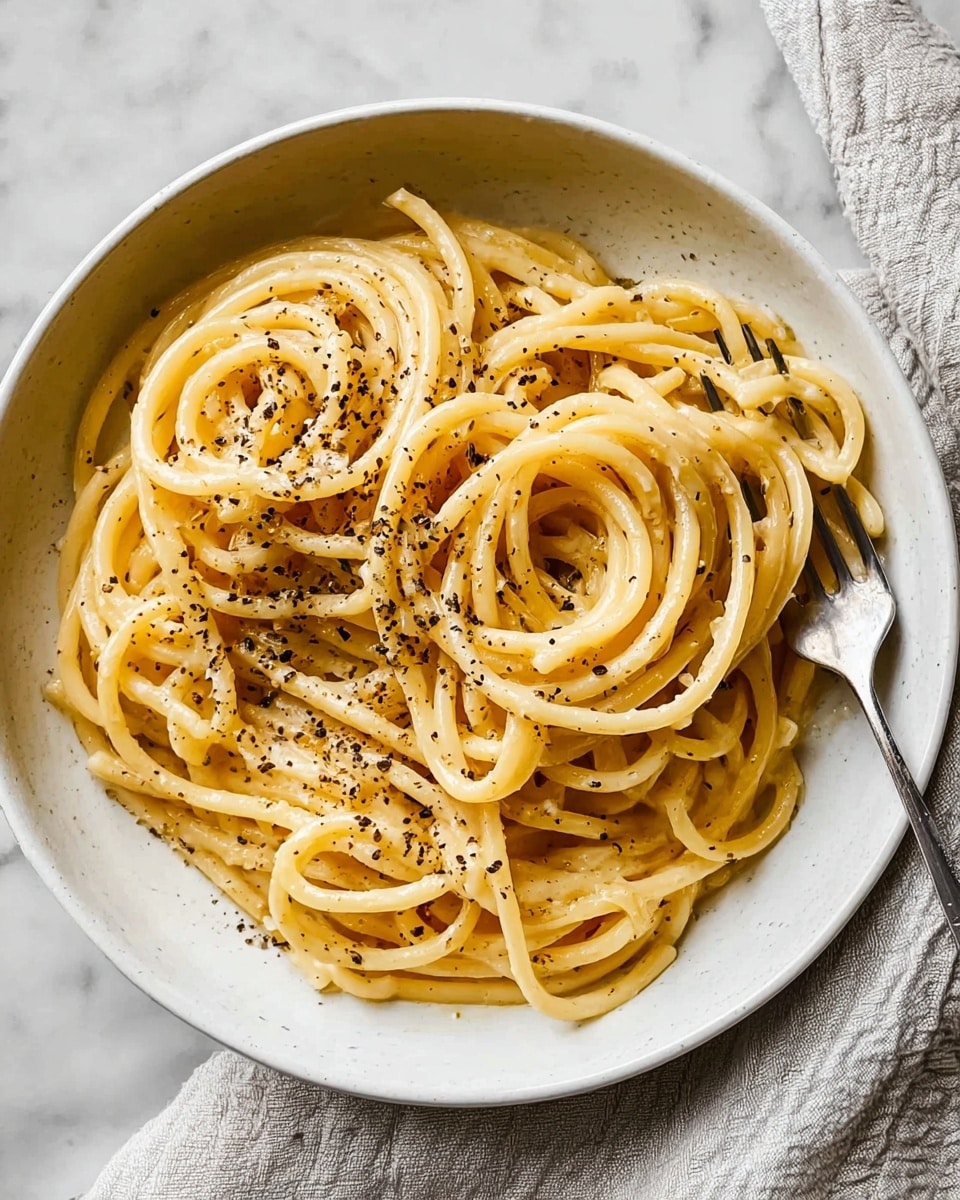 A white deep bowl holds a simple dish of bucatini pasta coated with a glossy light golden sauce, evenly mixed to give the noodles a smooth, slightly oily texture. The thick, hollow pasta strands are loosely twirled and piled up to form one main layer, with a generous sprinkle of cracked black pepper scattered on top, adding contrast with its dark specks. The bowl rests on a white marbled surface with a folded, light gray cloth and a metallic fork placed to the right side, creating a clean, minimalist setup. photo taken with an iphone --ar 4:5 --v 7