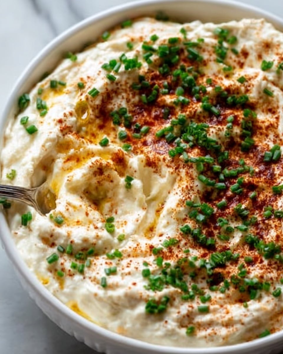 The image shows a round white bowl filled with a creamy, textured dip that has a light beige color. The top layer is sprinkled with a reddish-brown spice and finely chopped green herbs scattered evenly across the surface. A spoon rests inside the bowl, scooping out some of the creamy dip, giving a sense of softness and smoothness in the middle. The bowl is placed on a white marbled textured surface with a few pieces of light tan crackers visible in the background. photo taken with an iphone --ar 4:5 --v 7