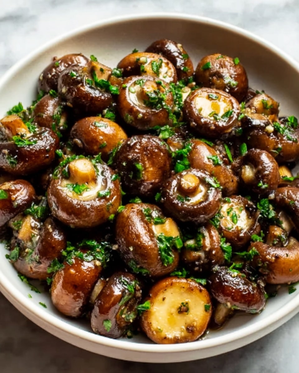 The image shows a white bowl filled with whole and halved brown mushrooms that are cooked to a shiny, golden brown. The mushrooms have a slightly glossy texture from oil or butter and are sprinkled with chopped green herbs and minced garlic, giving a fresh and vibrant touch. The mushrooms look plump and juicy, stacked closely together, with some small pieces of garlic visible on top. The bowl sits on a white marbled surface. Photo taken with an iphone --ar 4:5 --v 7