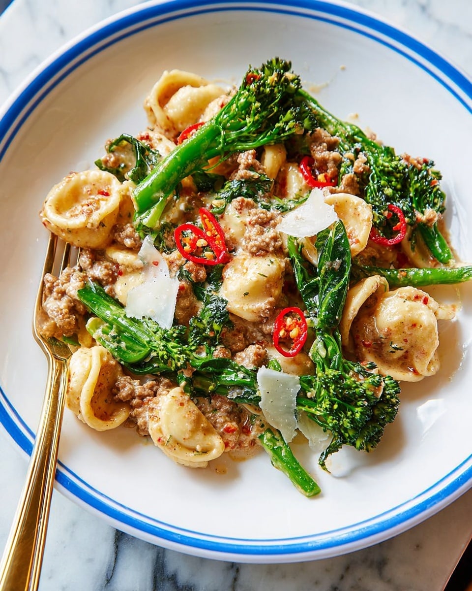 The image shows a white plate with blue rims filled with a colorful pasta dish. The base layer is small round orecchiette pasta, pale yellow in color, spread evenly across the plate. On top, there are bright green broccolini pieces with textured leafy tops and stalks scattered among the pasta. Red chili slices add pops of sharp red, contrasting with the green and yellow. The dish is topped with irregular thin shavings of pale white cheese, lightly scattered, along with bits of a creamy, lightly browned sauce that clings to both pasta and vegetables. A golden fork rests on the side, partially under the food. The background is a white marbled texture. photo taken with an iphone --ar 4:5 --v 7