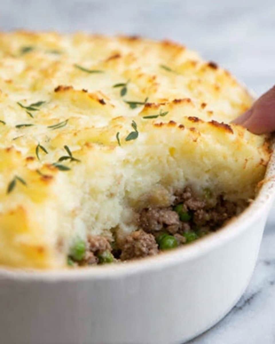 The image shows a white round baking dish filled with a shepherd's pie. The bottom layer is dark brown cooked ground meat mixed with small green peas. On top is a thick, smooth layer of mashed potatoes, which are light in color with some golden brown spots where the surface was baked. Small thin green herb pieces are scattered on top as garnish. The dish is set on a white marbled surface. photo taken with an iphone --ar 4:5 --v 7