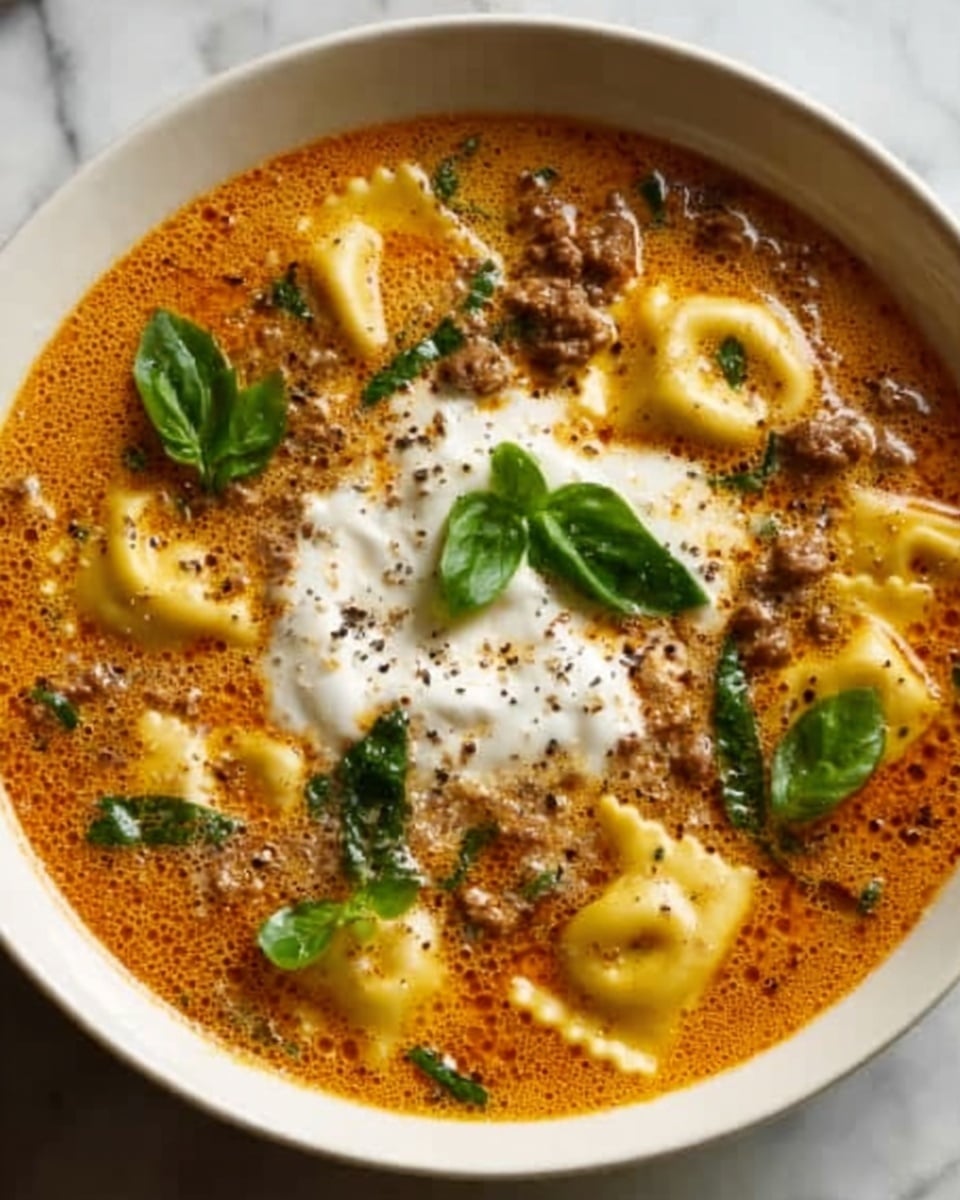 A white bowl filled with a rich, creamy tomato soup topped with small ravioli pieces scattered throughout the soup. The soup itself is a deep reddish-orange color with a textured surface showing herbs and specks of ground black pepper. In the center, there is a dollop of white cream, slightly melting into the soup, garnished with fresh green basil leaves. The edges of the ravioli have a light yellow color with ruffled edges, giving a soft texture contrast against the smooth soup. The bowl is set on a white marbled surface. photo taken with an iphone --ar 4:5 --v 7