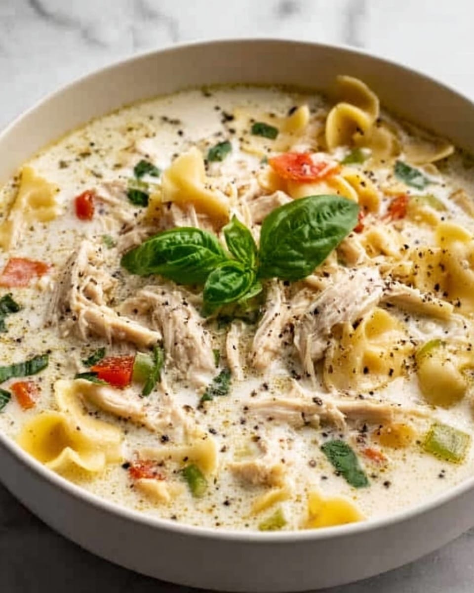 This image shows a white bowl filled with creamy chicken and tortellini soup. The soup has a thick white cream base with visible torn pieces of shredded chicken scattered on top. Yellow tortellini pasta floats through the creamy broth, some partially submerged, showing their folded, round shapes. There are small bits of orange carrot and green spinach or basil leaves spread evenly across the surface. Black pepper is sprinkled over the soup, adding contrast to the light colors. A single fresh green basil leaf is placed in the center as a garnish. The bowl sits on a white marbled surface. Photo taken with an iphone --ar 4:5 --v 7