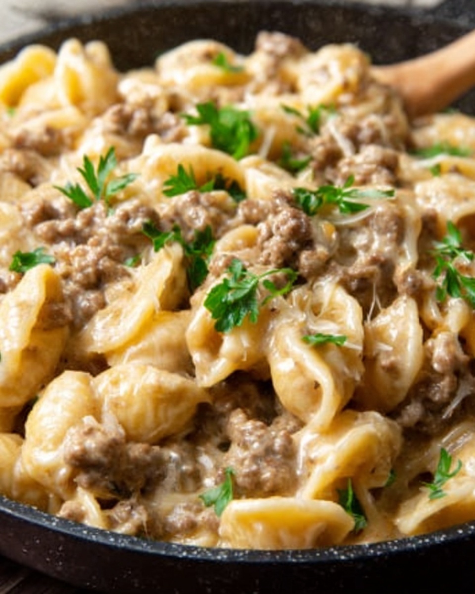 The image shows a close-up of a creamy pasta dish in a black pan. The pasta is shell-shaped, coated with a thick, beige creamy sauce mixed with small pieces of cooked brown ground meat. On top, there are some bright green parsley leaves scattered as garnish. The sauce looks smooth and rich, covering the pasta well, and the meat pieces are evenly spread throughout. The photograph is detailed, highlighting the texture of the sauce and the freshness of the parsley. The photo taken with an iphone --ar 4:5 --v 7