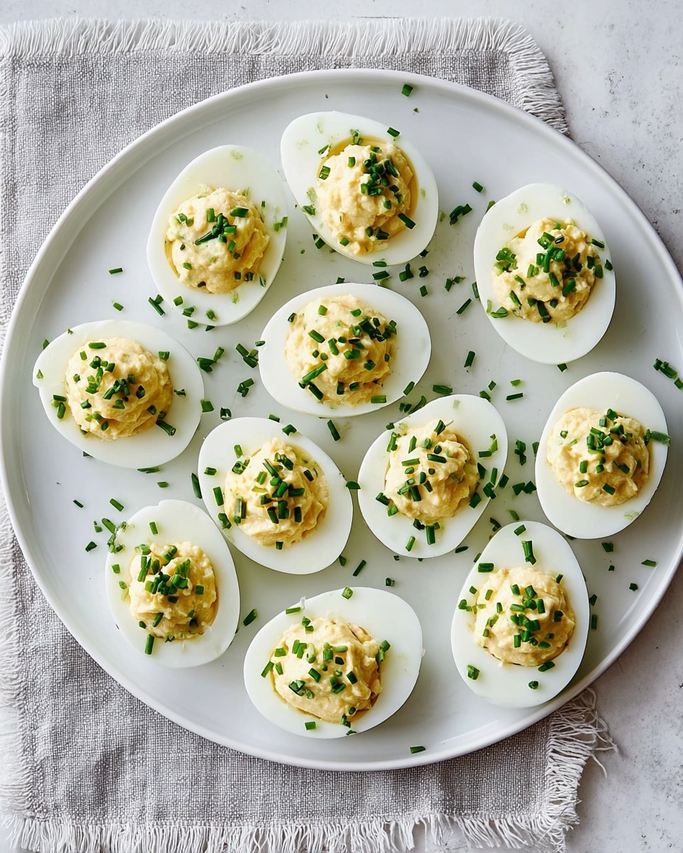 A white round plate holds thirteen half boiled eggs arranged randomly, each egg has two layers: the bottom white layer is smooth and shiny egg white, and the top layer is a creamy, pale yellow egg yolk mixture with a slightly chunky texture, sprinkled with small pieces of bright green chopped chives. The plate rests on a white marbled textured surface with a white cloth partially visible on the right side. photo taken with an iphone --ar 4:5 --v 7