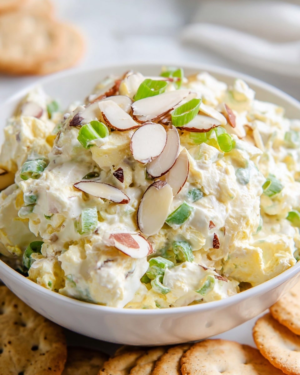 A white bowl filled with a creamy, chunky mixture that has visible layers of pale yellow cheese, white creamy dressing, thinly sliced green onions, and sliced almonds scattered throughout. The salad looks thick and textured with small, soft curds and crunchy pieces mixed evenly. Behind the bowl, there are light brown crackers partially in focus, resting on a white marbled surface. photo taken with an iphone --ar 4:5 --v 7