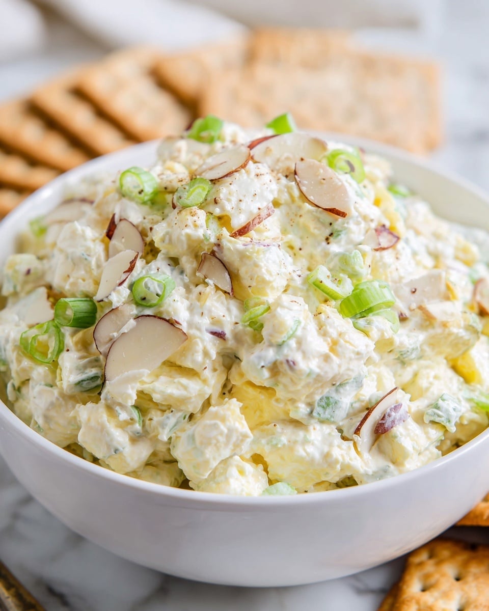 A close-up of a white bowl filled with a creamy salad mixture consisting of roughly three layers: the base layer is a soft, chunky, pale yellow mixture, likely a cheese or egg salad base; mixed in are bright green chopped scallions scattered throughout; and thin, oval slices of almonds with a light brown edge and white center sit on the top and mixed inside, adding texture. The white bowl rests on a white marbled surface with beige crackers in the background. The lighting is bright and natural, highlighting the creamy texture of the salad, with sharp detail on the individual almond slices and scallions. Photo taken with an iphone --ar 4:5 --v 7